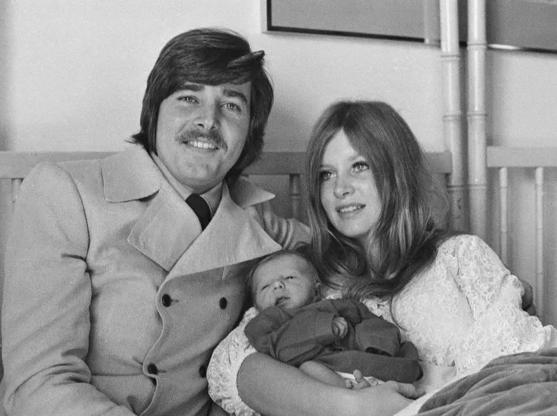 بوبي شيرمان يجلس مع زوجته وابنهما الرضيع، مبتسمين في لحظة عائلية دافئة، تعكس حياته كفنان مشهور في الستينيات.