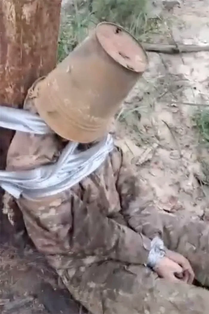 جندي روسي مربوط إلى شجرة مع دلو على رأسه، يُظهر مشهدًا من سوء المعاملة في ساحة المعركة، وسط توتر الحرب الأوكرانية.