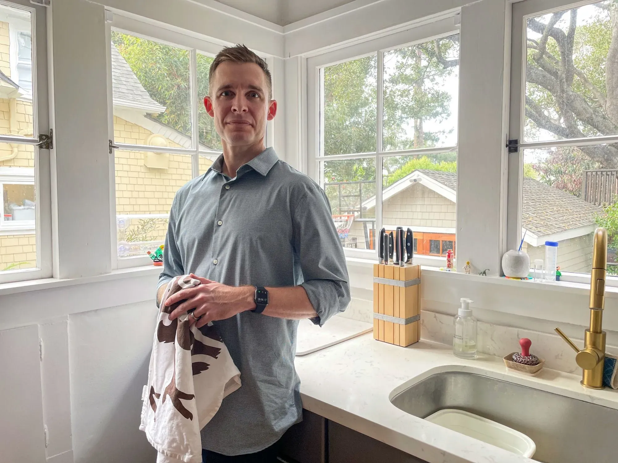 رجل يقف في المطبخ ممسكًا بمنشفة، يبدو أنه يشارك في الأعمال المنزلية. تعكس الصورة التحديات المتعلقة بتوزيع الأعباء المنزلية بين الأزواج.