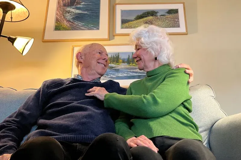 صورة لزوجين مسنين يجلسان معًا على أريكة، يتبادلان الابتسامات واللمسات الحانية، مع لوحات طبيعية معلقة خلفهما، تعكس الحب الدائم والتواصل العاطفي.