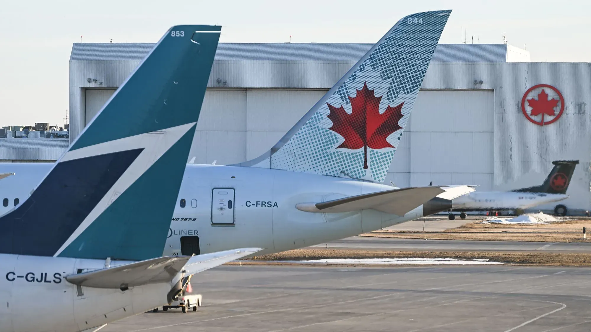ذيل طائرتين تجاريتين في مطار، حيث يظهر شعار شركة الطيران الكندية وعلم كندا، مع خلفية لمرافق المطار.