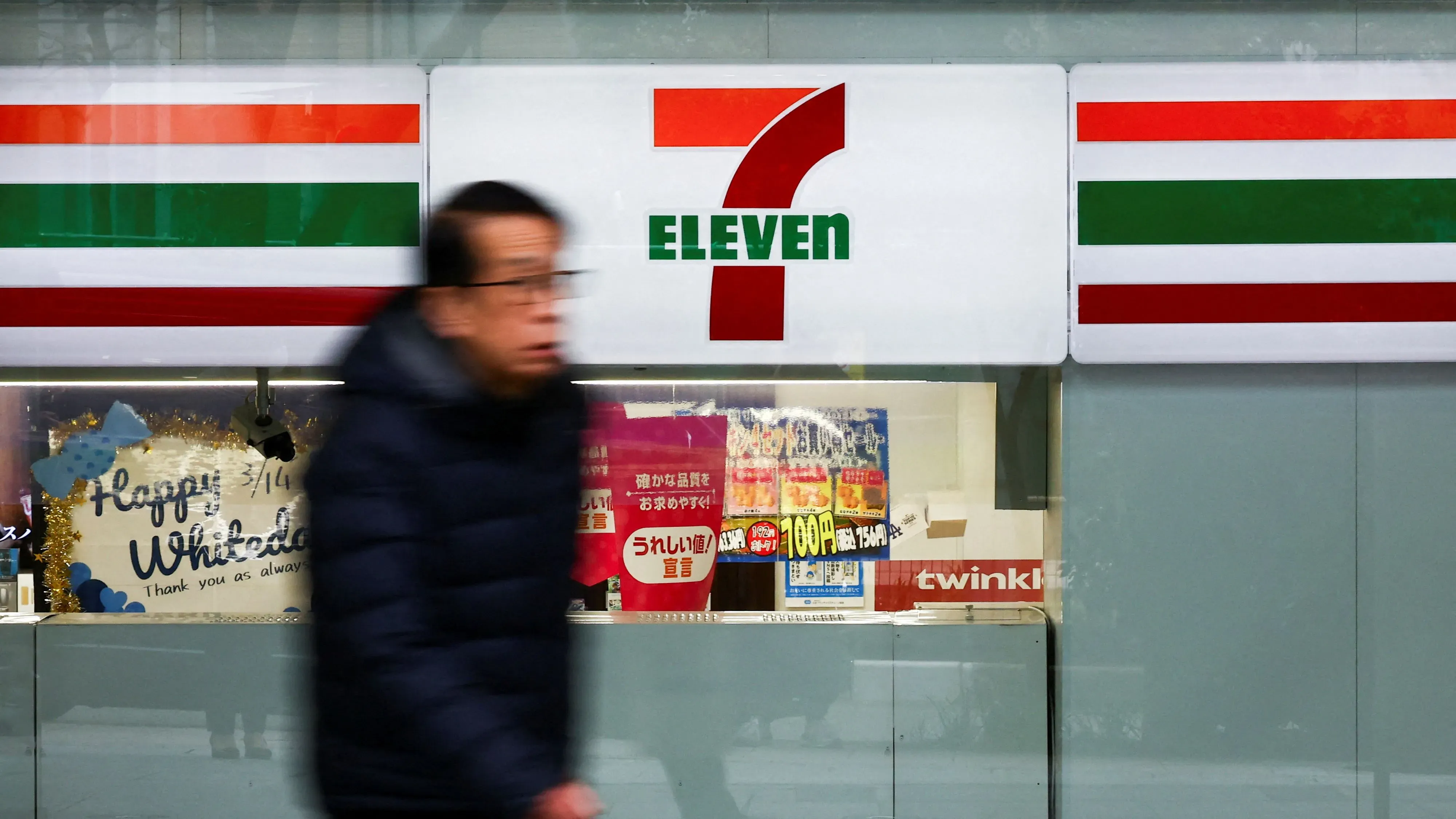 رجل يمر بجوار واجهة متجر 7-Eleven في اليابان، حيث يظهر الشعار واللافتات المحيطة، مع لافتات ترويجية في الخلفية.