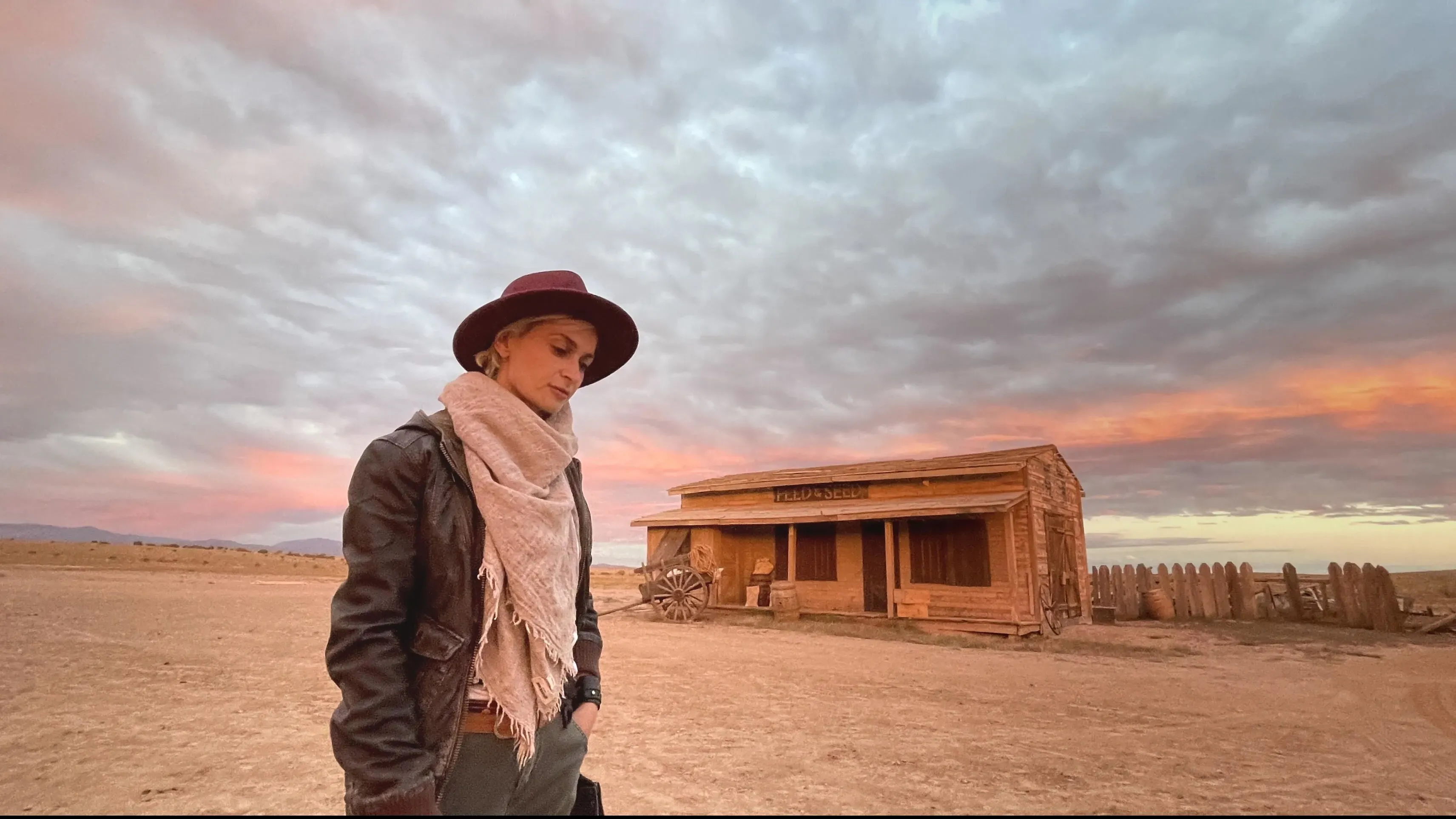 امرأة ترتدي قبعة وحجاباً، واقفة في موقع تصوير فيلم "Rust" أمام مبنى خشبي، مع سماء ملونة بالخيوط الوردية والبرتقالية.