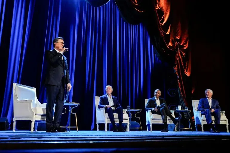 ستيفن كولبير يتحدث على المسرح أمام ثلاثة ضيوف في حدث خاص، مع خلفية من الستائر الزرقاء، يعكس أجواء برنامج حواري.