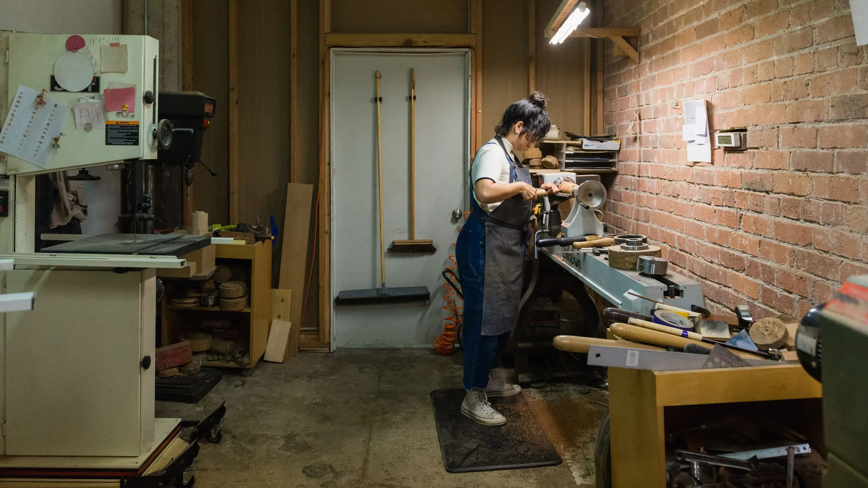 امرأة تعمل في ورشة صغيرة، تستخدم أدوات الحرف اليدوية لتصميم منتجات، وسط بيئة من الطوب المكشوف وأثاث بسيط.