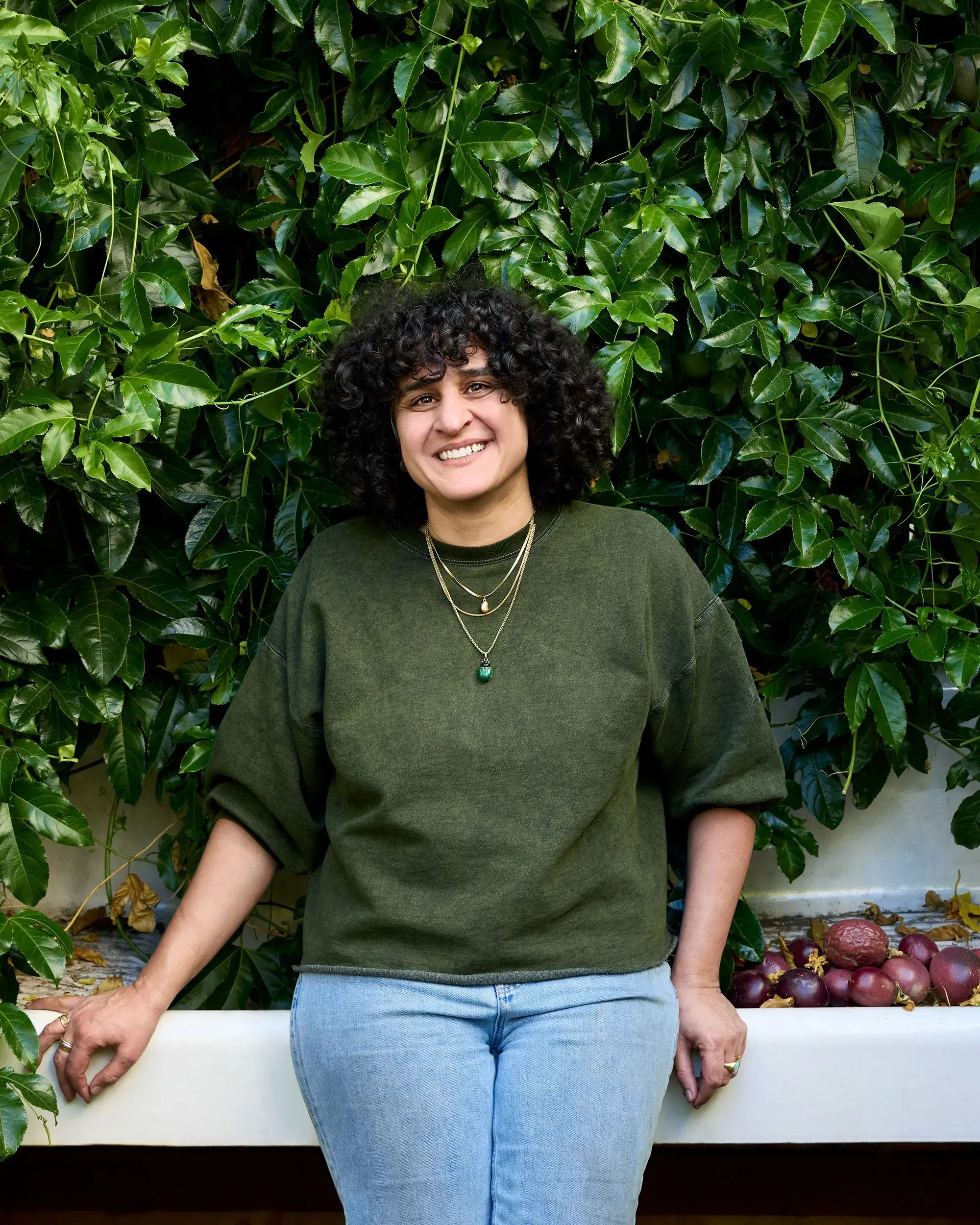 شخصية مبتسمة ترتدي سترة خضراء، تقف أمام حائط مغطى بأوراق خضراء، مع ثمار فاكهة خلفها، تعكس أجواء مريحة ومحببة.