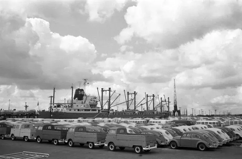 صورة تظهر مجموعة من الشاحنات الصغيرة والسيارات في ميناء، مع سفينة شحن كبيرة في الخلفية. تعكس الصورة تأثير التعريفات الجمركية على صناعة السيارات.