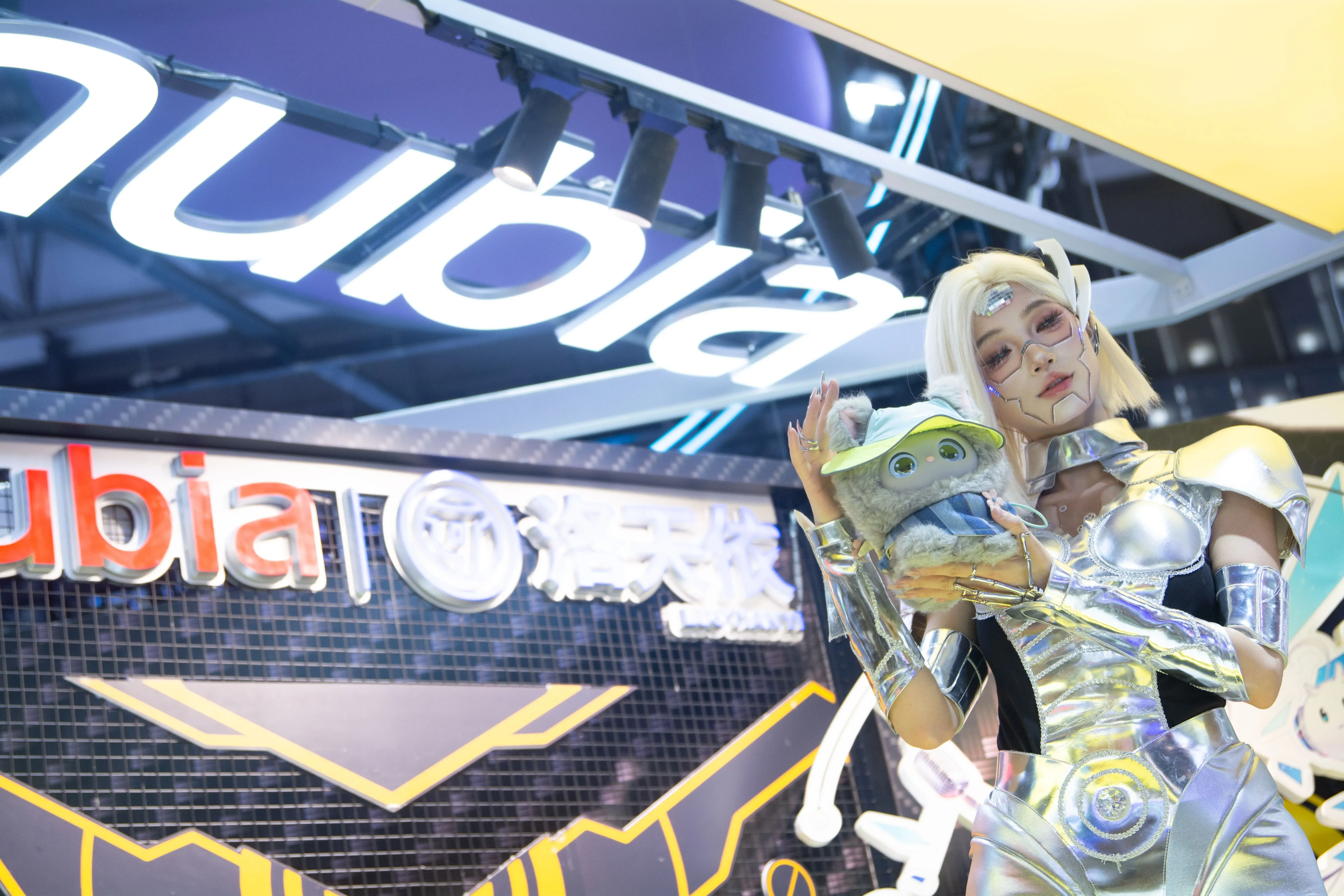 عارضة ترتدي زيًا فضائيًا لامعًا، تحمل دمية تمثل شخصية خيالية، أمام شعار شركة "نوبيا" في مؤتمر الذكاء الاصطناعي.