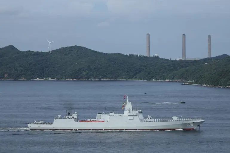 سفينة حربية صينية تبحر في المياه المحيطة بتايوان، مع ظهور التلال والمرافق الصناعية في الخلفية، في إطار تدريبات عسكرية.