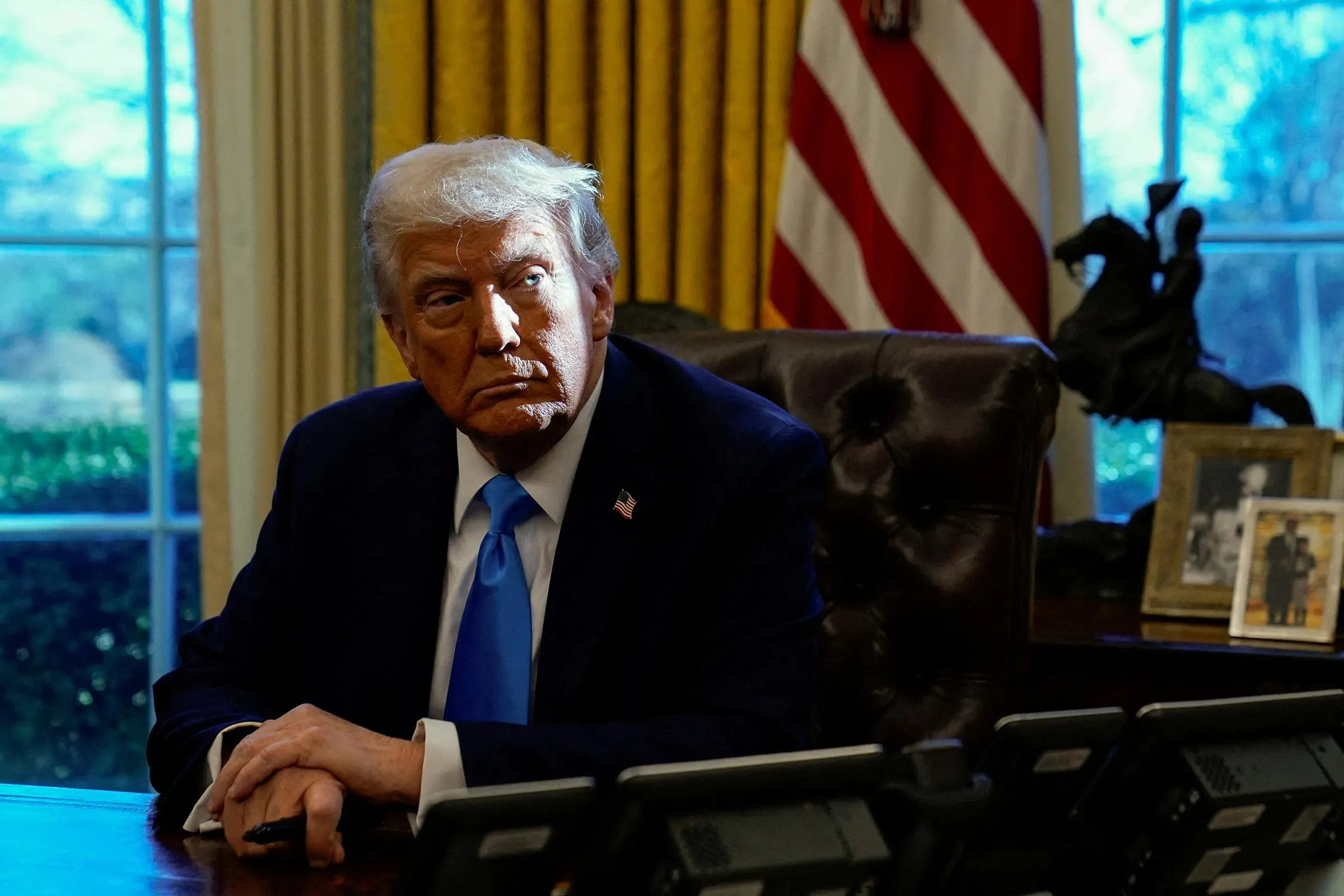 ترامب يجلس في المكتب البيضاوي، مع تعبير وجهه الجاد، محاطًا بأعلام أمريكية وصور عائلية، يعكس مشاعر القلق حول أداءه الاقتصادي.