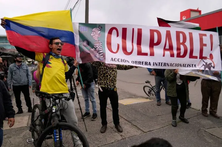محتجون يحملون لافتة مكتوب عليها "CULPABLE!" ويرتدون ملابس ملونة، مع العلم الكولومبي، في تظاهرة ضد الرئيس السابق ألفارو أوريبي.