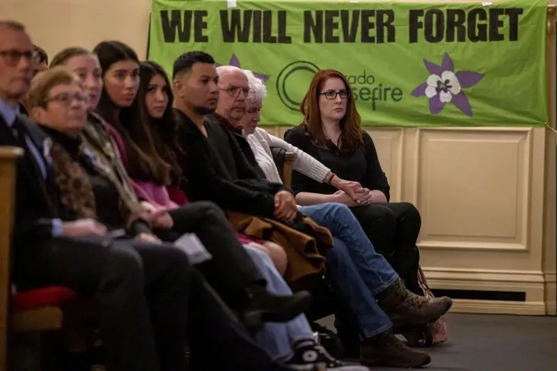 مجموعة من الأشخاص يجلسون في حدث تأبيني، مع لافتة خلفهم تحمل عبارة "لن ننسى أبدًا"، تعبيرًا عن الذكرى والتضامن بعد مأساة كولومباين.