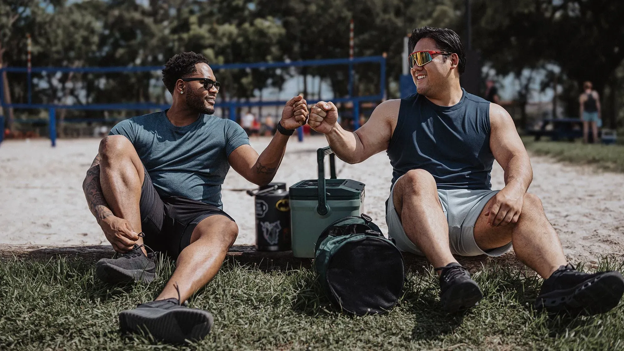 رجلان يجلسان معًا على العشب، يتبادلان التحية بقبضة اليد، يجسدون روح الصداقة والتواصل بين الرجال في بيئة غير رسمية.