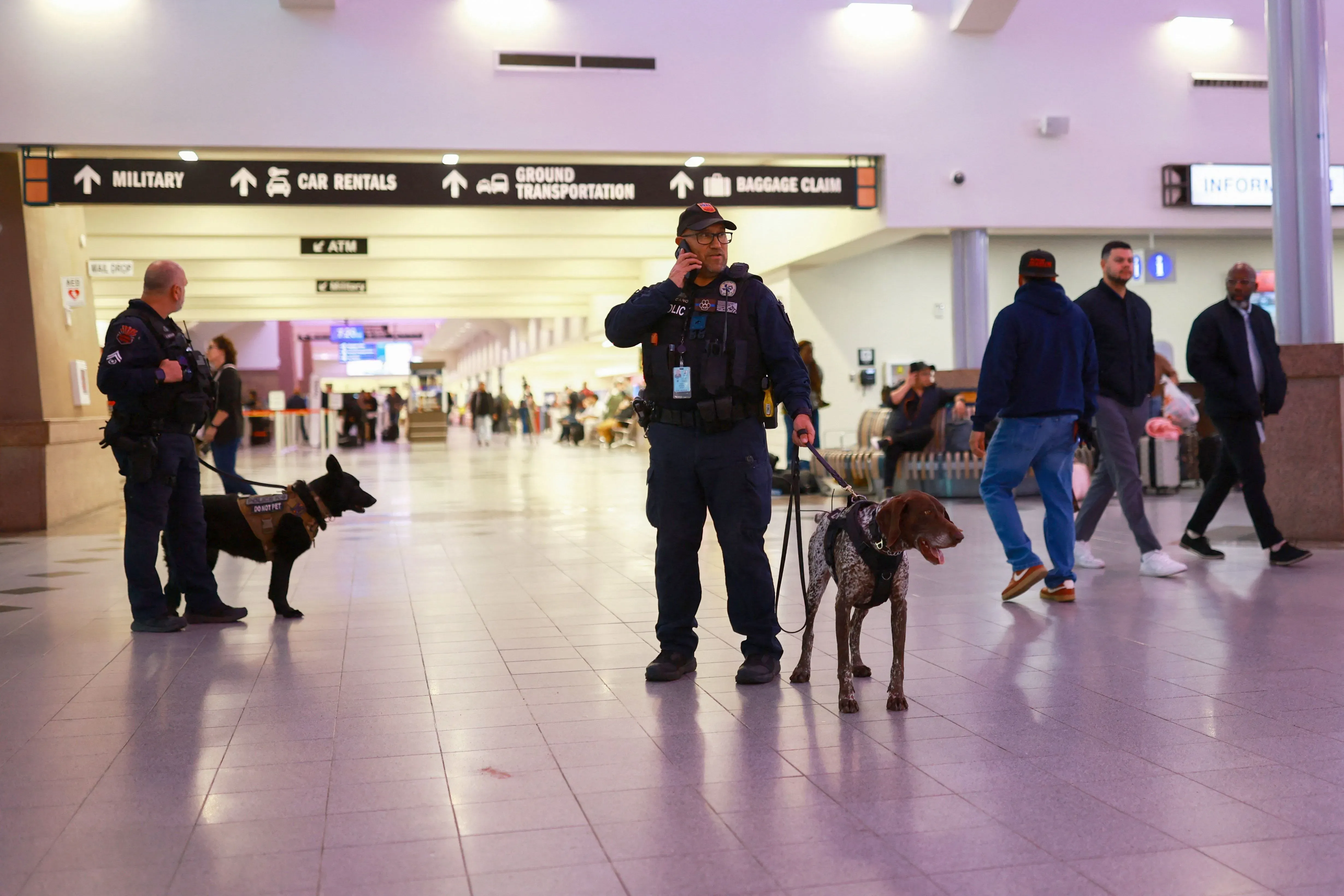 رجال أمن يرتدون زيًا رسميًا في مطار، مع كلاب بوليسية، يتفقدون المنطقة وسط حركة المسافرين، في سياق إغلاق المجال الجوي.