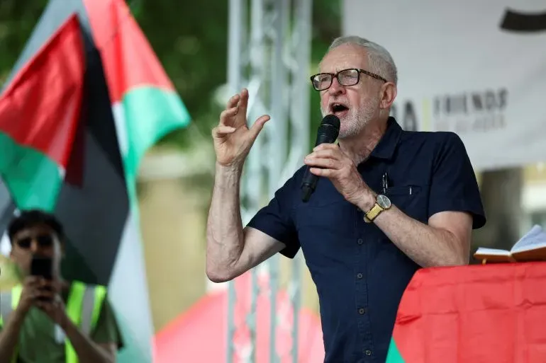 جيريمي كوربين يتحدث في تجمع سياسي، محاطًا بأعلام فلسطينية، مع التركيز على قضايا العدالة الاجتماعية والسياسية.