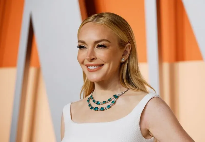 ليندسي لوهان مبتسمة في حدث، ترتدي فستاناً أبيض مع قلادة مميزة. تعكس إطلالتها التزامها بالمظهر الشاب والجذاب.