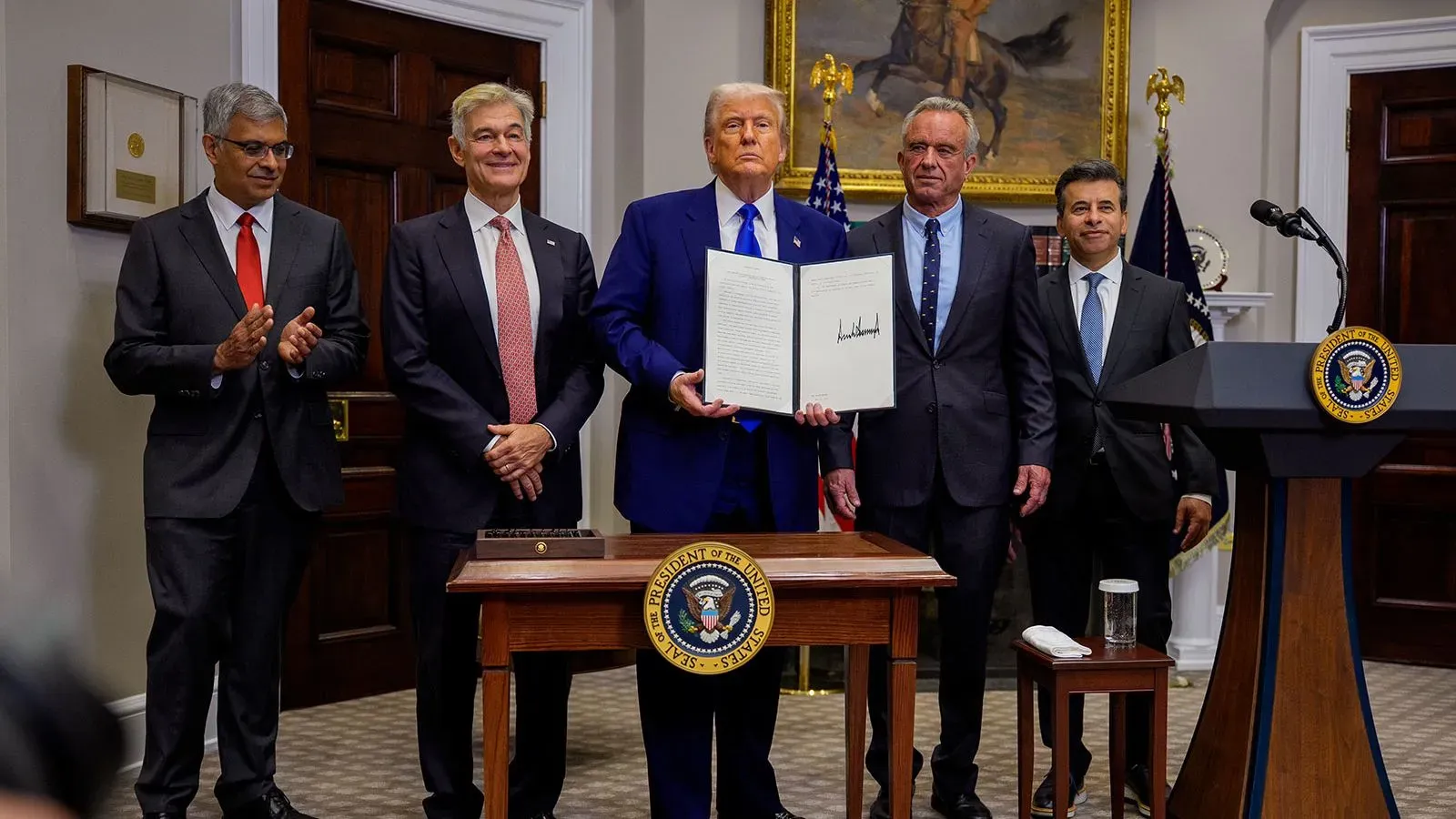 اجتماع في البيت الأبيض حيث يقف ترامب مع مجموعة من المستشارين، يحمل وثيقة رسمية، بينما يحيط به أربعة رجال في قاعة رسمية.