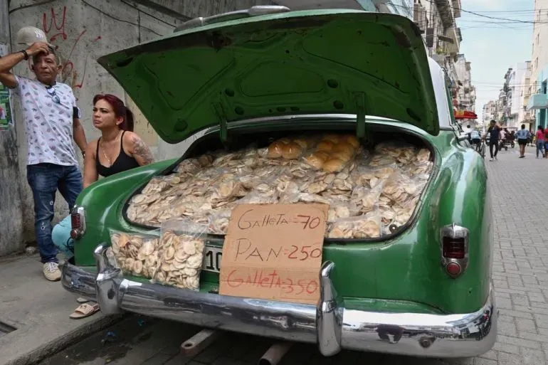 سيارة خضراء قديمة تحمل كميات كبيرة من الخبز والمعجنات في صندوقها الخلفي، مع وجود لافتة توضح الأسعار، في مشهد يعكس الأوضاع الاقتصادية في كوبا.