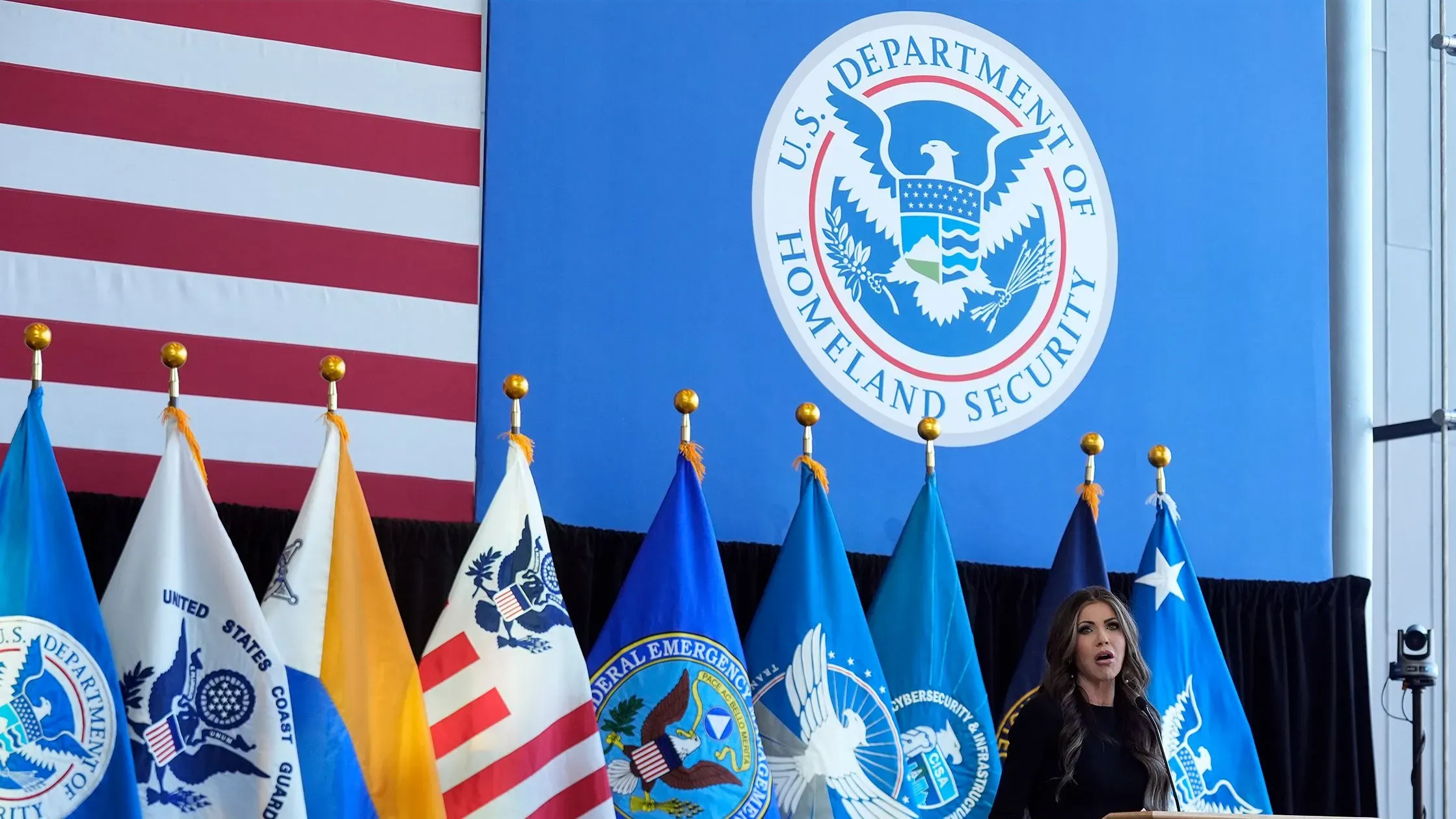 وزيرة الأمن الداخلي كريستي نويم تتحدث في مؤتمر صحفي، مع علم الولايات المتحدة وشعارات الوكالات الحكومية خلفها، وسط قلق حول الرقابة على احتجاز المهاجرين.
