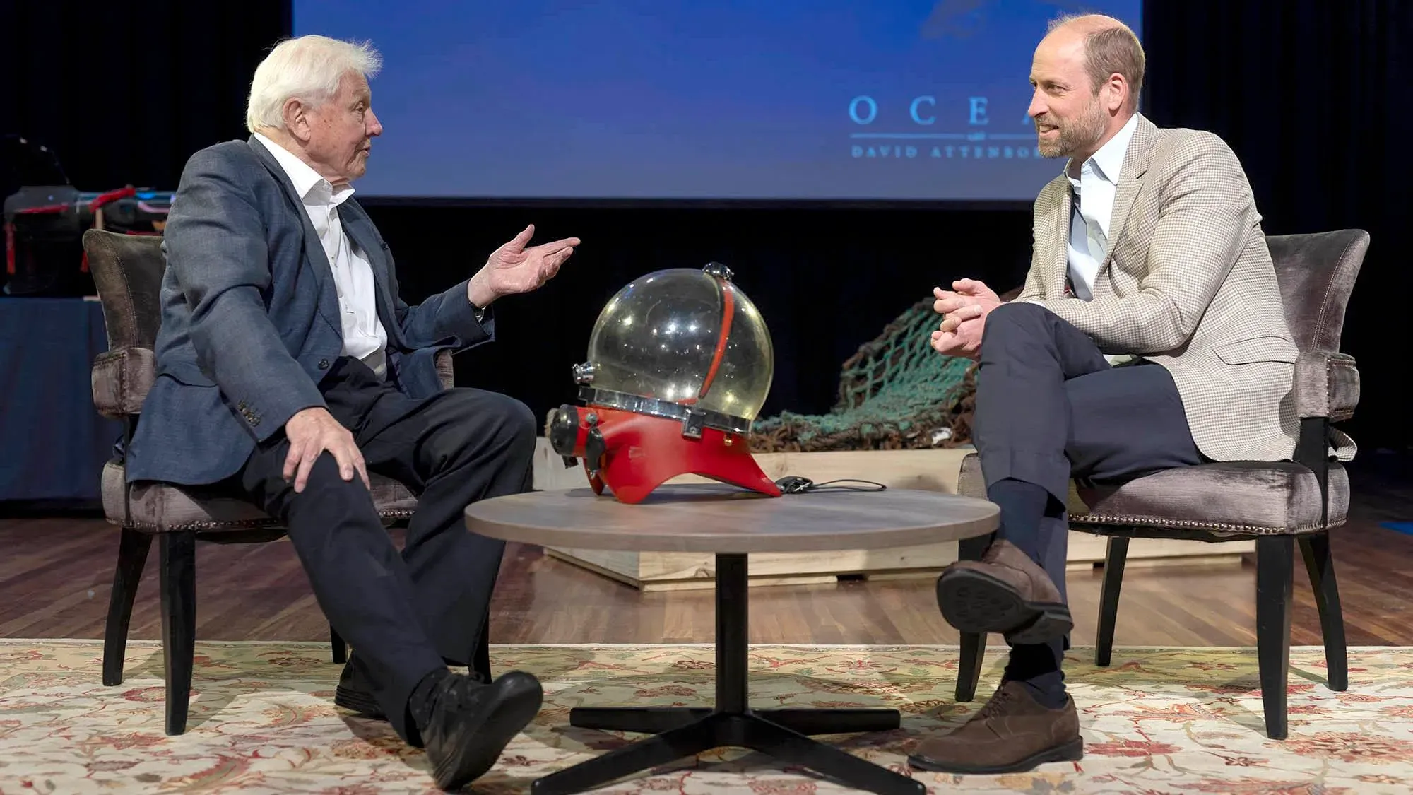 ديفيد أتينبورو والأمير ويليام يتحدثان عن تأثير الإنسان على المحيطات، مع وجود خوذة غوص حمراء على الطاولة.