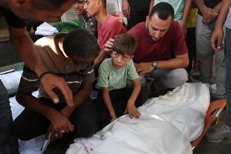 طفل صغير يبكي بجوار جثمان مغطى، محاط بأشخاص يعبرون عن حزنهم. تعكس الصورة معاناة المدنيين في غزة جراء الحرب.