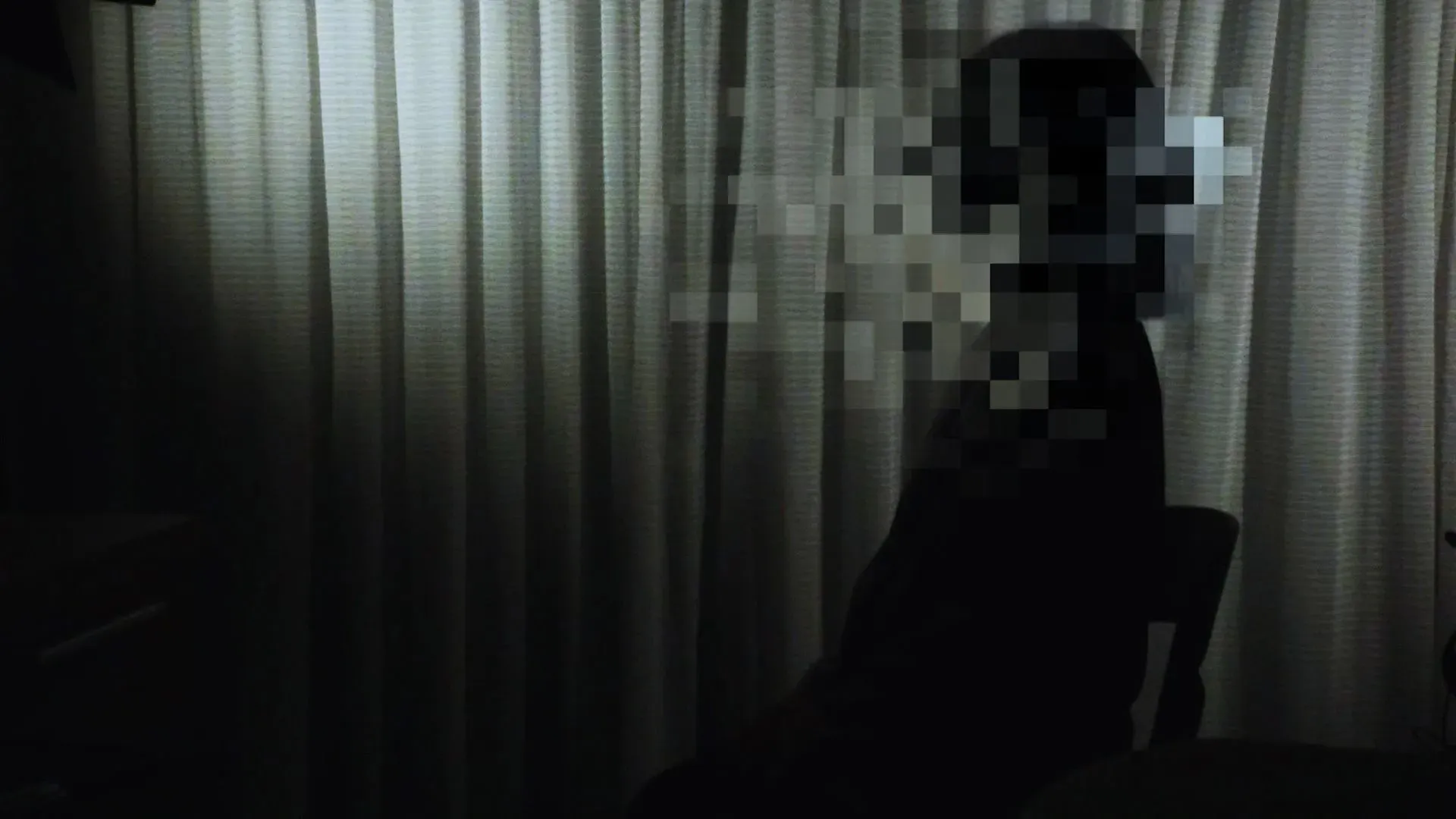 صورة ظلية لشخص يجلس في غرفة مظلمة، مع ستائر خلفه، تعكس شعور العزلة والقلق المرتبط بالجرائم الجنسية الرقمية.