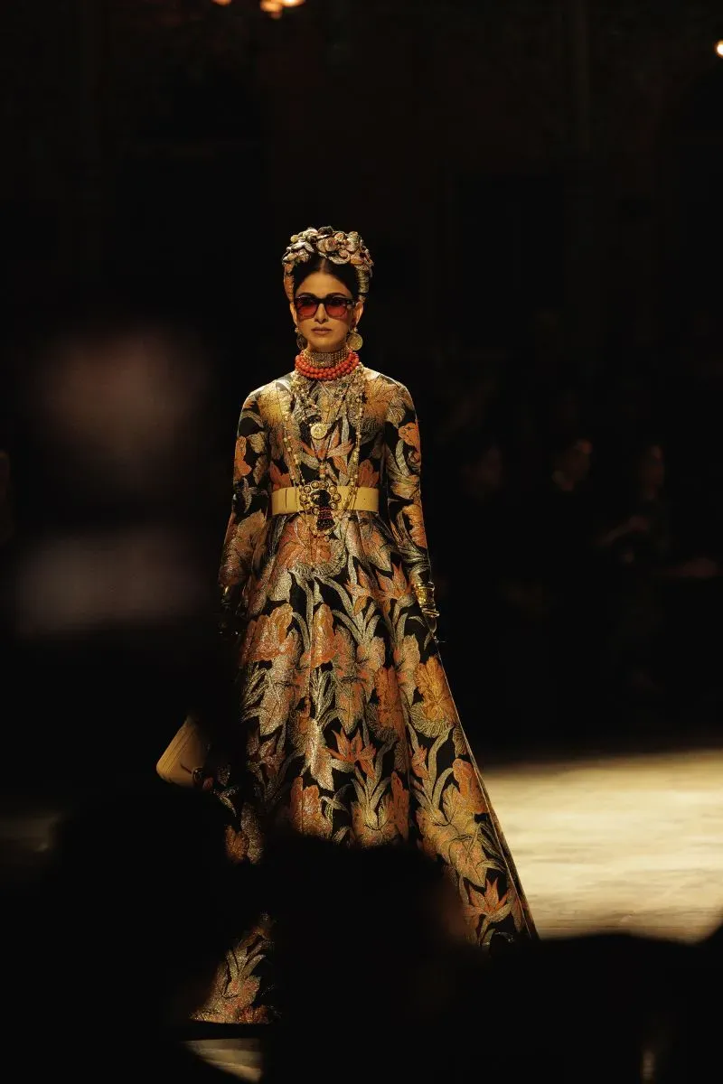 عارضة أزياء تسير بثقة على المسرح خلال عرض سابياساتشي موخرجي، مرتدية فستانًا مزخرفًا بألوان دافئة مع إكسسوارات مبهرة، وسط أضواء خافتة.