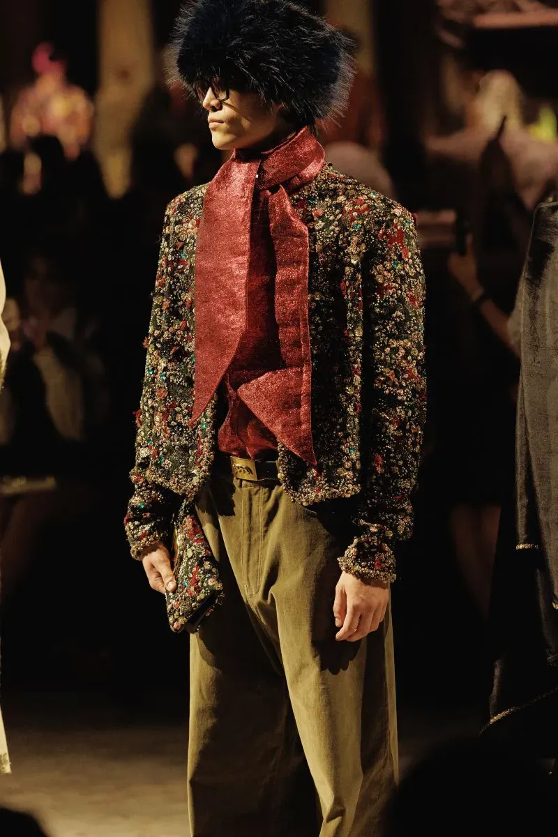 عارض أزياء يرتدي سترة مزخرفة بألوان متعددة مع وشاح أحمر وسروال أخضر داكن في عرض أزياء سابياساتشي موخرجي، يعكس أسلوبه الفريد والمبتكر.