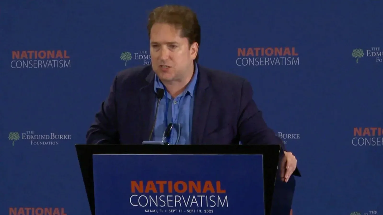 دارين بيتي يتحدث في مؤتمر "التحفظ الوطني" في ميامي، مع خلفية تتضمن شعار المؤتمر، مما يبرز دوره كمسؤول في وزارة الخارجية.