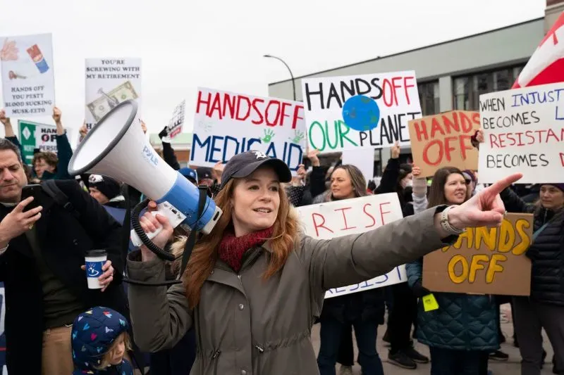 امرأة تحمل ميكروفونًا خلال احتجاج، محاطة بالمتظاهرين الذين يحملون لافتات تدعو إلى حماية البيئة والرعاية الصحية.
