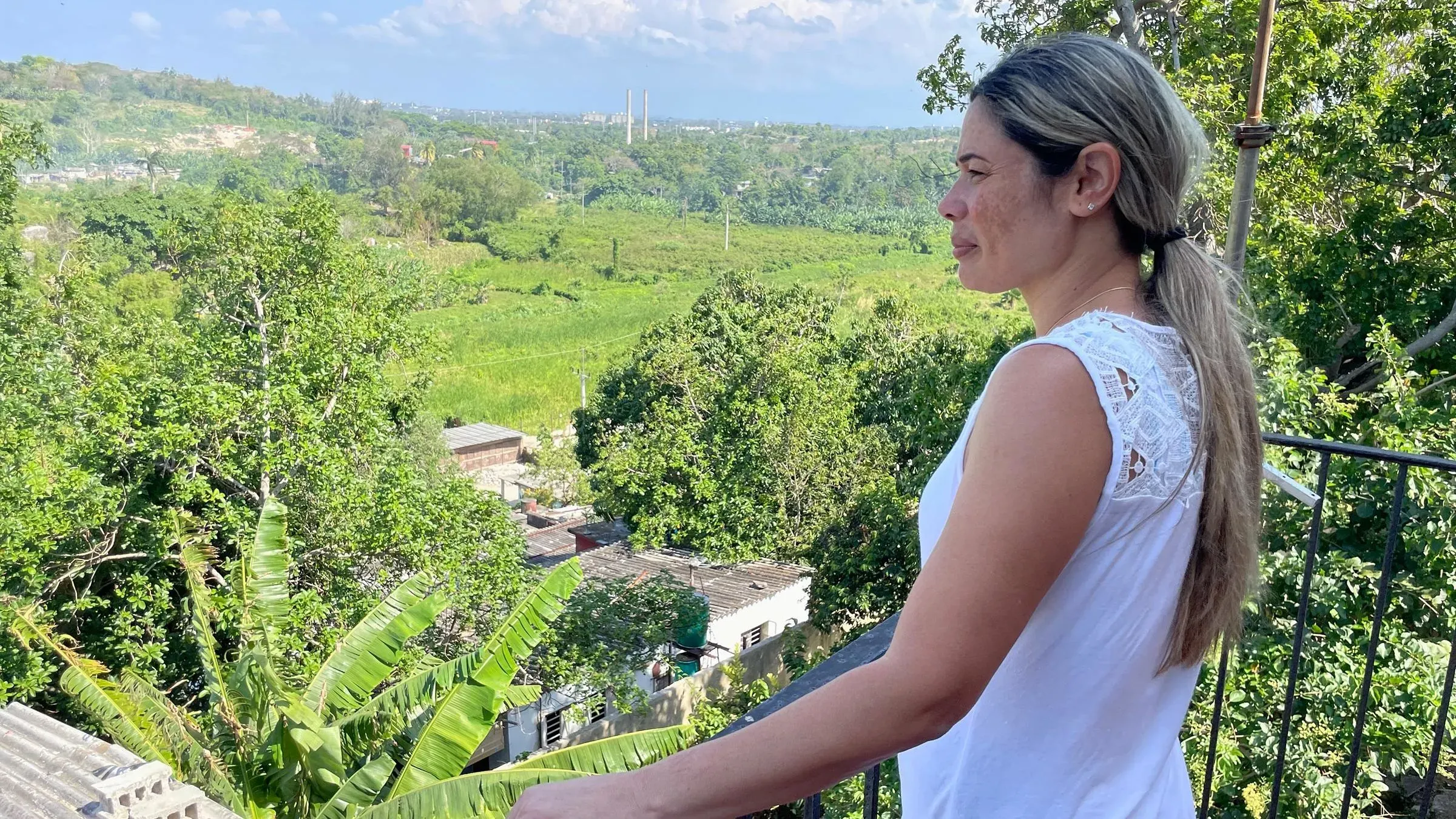 هايدي سانشيز تقف في شرفة منزلها المتهدم في كوبا، تنظر إلى المناظر الطبيعية الخضراء، تعكس مشاعر التحدي والأمل بعد الترحيل.