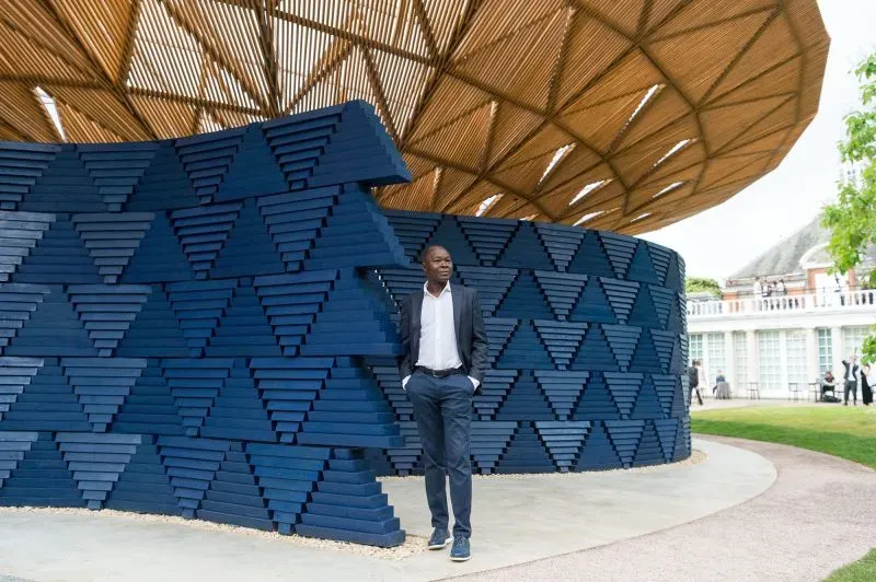 جناح سربنتين المعماري بتصميمه الفريد، يتكون من خشب ومثلثات زرقاء، مع شخص يقف بجانبه في حدائق كنسينغتون بلندن.
