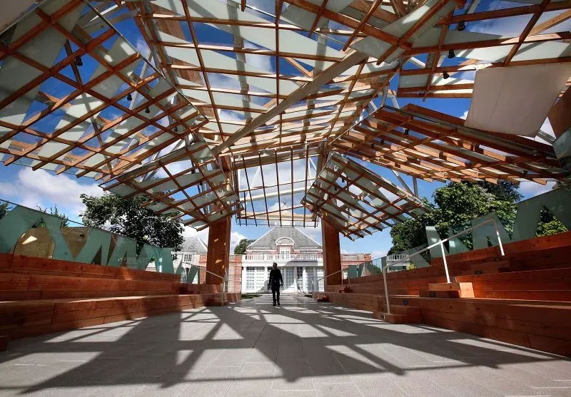 جناح سربنتين المعماري بتصميمه الفريد، يجمع بين الخشب والزجاج، مع شجرة جينكو في الوسط، يعكس تأثير الضوء في حدائق كنسينغتون.