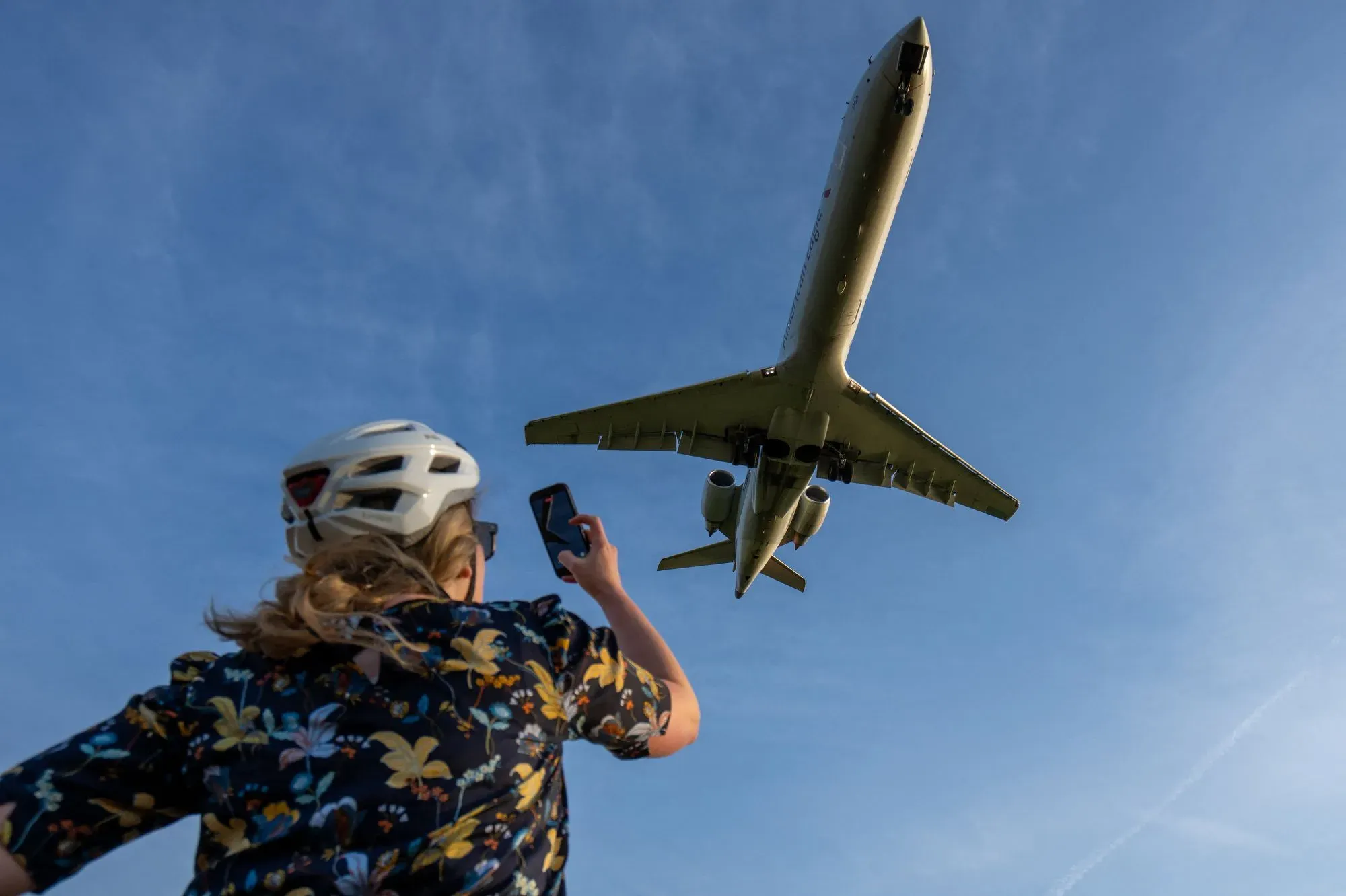 امرأة ترتدي خوذة تلتقط صورة لطائرة تحلق في السماء، مما يعكس تأثير ارتفاع أسعار وقود الطائرات على السفر الجوي.