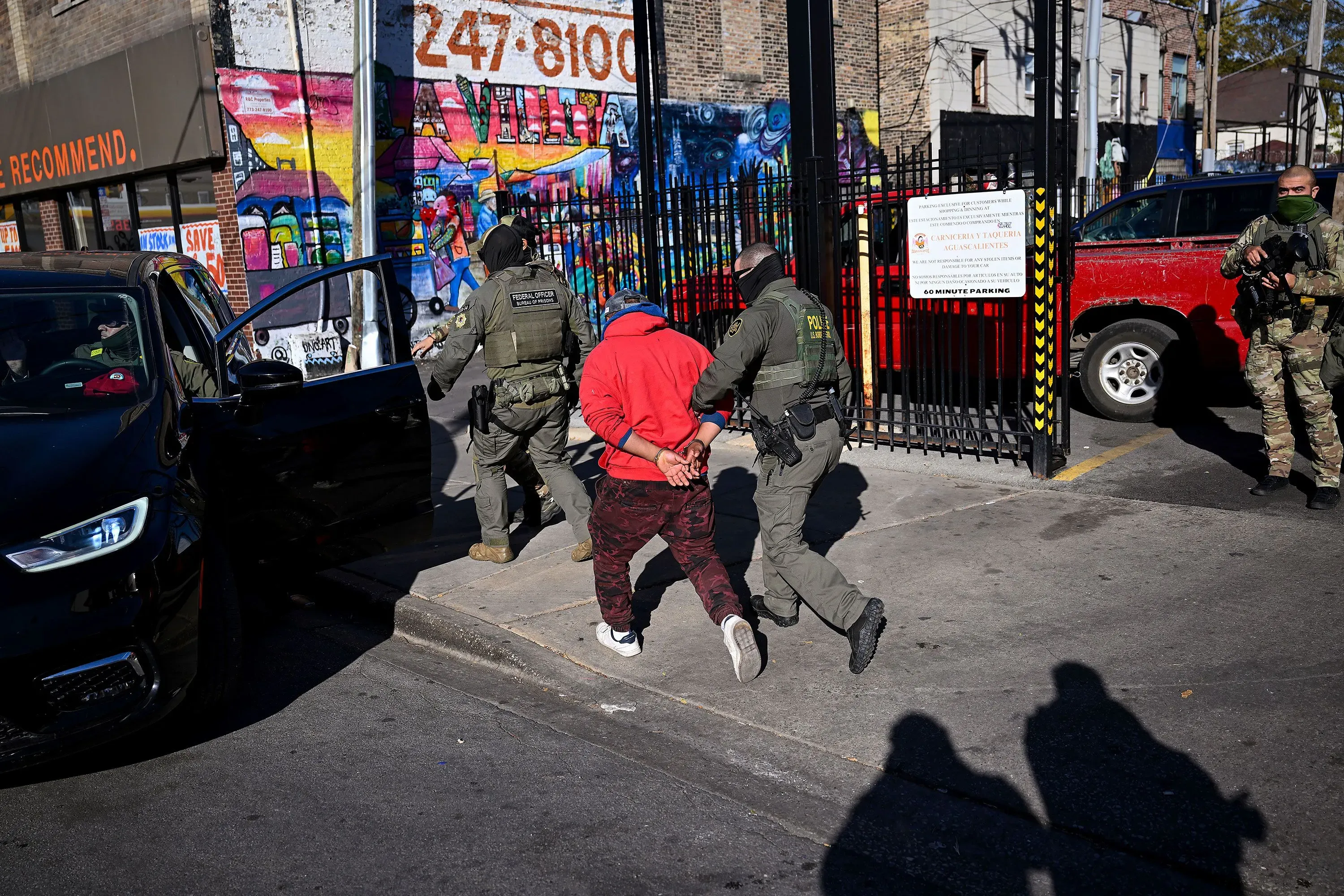 اعتقال شخص يرتدي سترة حمراء من قبل ضباط الهجرة في منطقة حضرية، مع جدارية ملونة خلفهم، مما يعكس التوترات المتعلقة بالهجرة.