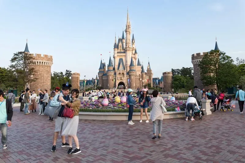 منتجع ديزني لاند أبوظبي الجديد يظهر قلعة خيالية مهيبة تحيط بها أزهار ملونة وزوار يتجولون في أجواء احتفالية.