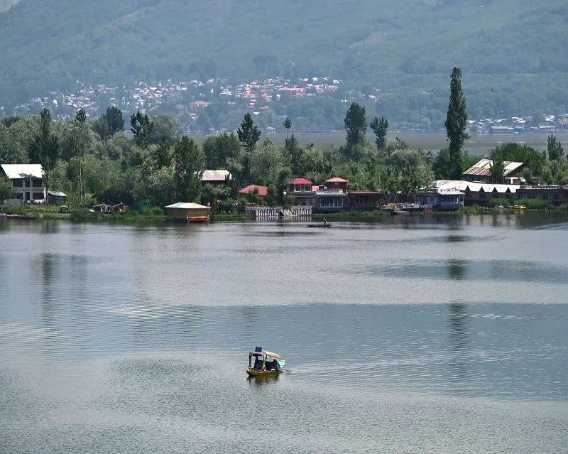 منظر طبيعي في كشمير يظهر بحيرة هادئة محاطة بأشجار الصنوبر ومنازل تقليدية، تعكس جمال المنطقة المتنازع عليها.