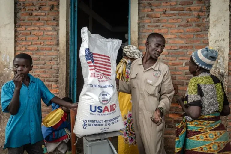 توزيع مساعدات غذائية من الوكالة الأمريكية للتنمية الدولية (USAID) في رواندا، حيث يتحدث رجل مع امرأتين بجانب كيس من المساعدات.