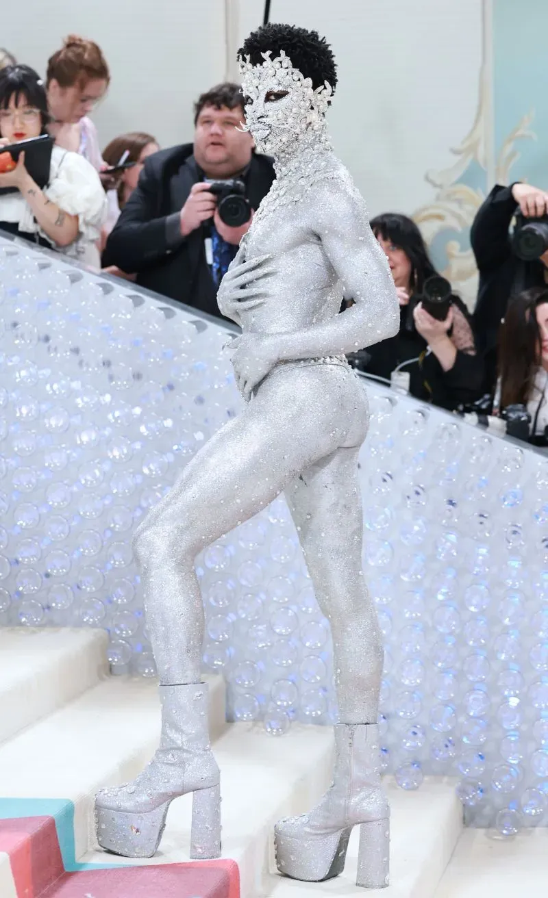 يظهر شخص يرتدي زيًا فضيًا لامعًا وأحذية بكعب عالٍ في حدث للأزياء، محاطًا بمصورين. تعكس الإطلالة تحديًا للمعايير التقليدية للجنسين.