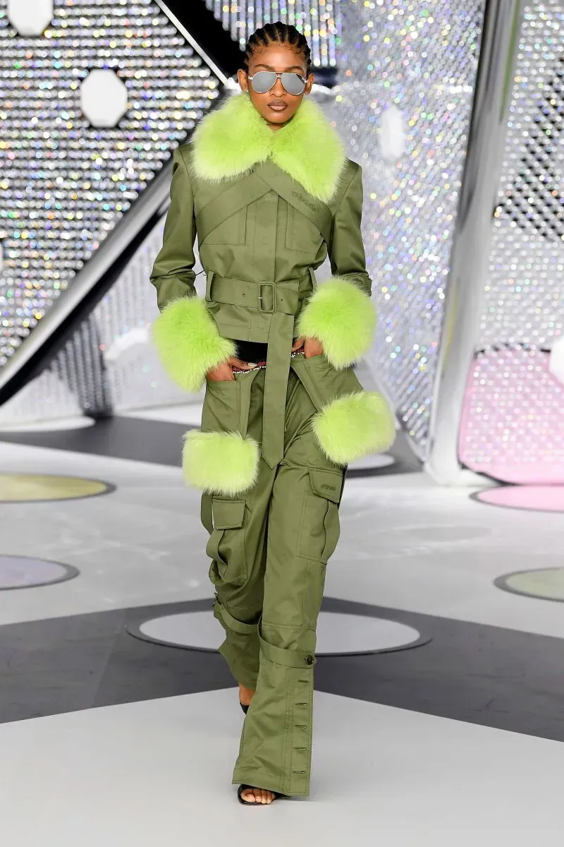 عارضة أزياء ترتدي سترة خضراء مع ياقة من الفرو اللامع، وتعرض تصميمًا عصريًا يتضمن جيوبًا كبيرة، خلال عرض أزياء.