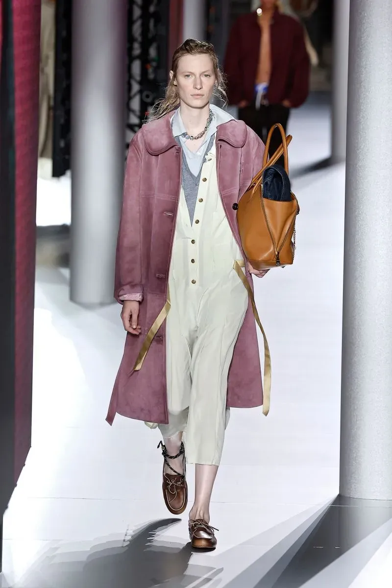 عارضة أزياء ترتدي معطفًا ورديًا طويلًا مع بنطال أبيض، تحمل حقيبة جلدية بنية، تسير في عرض أزياء يعكس أسلوب "المرأة الإنجليزية المرهقة".