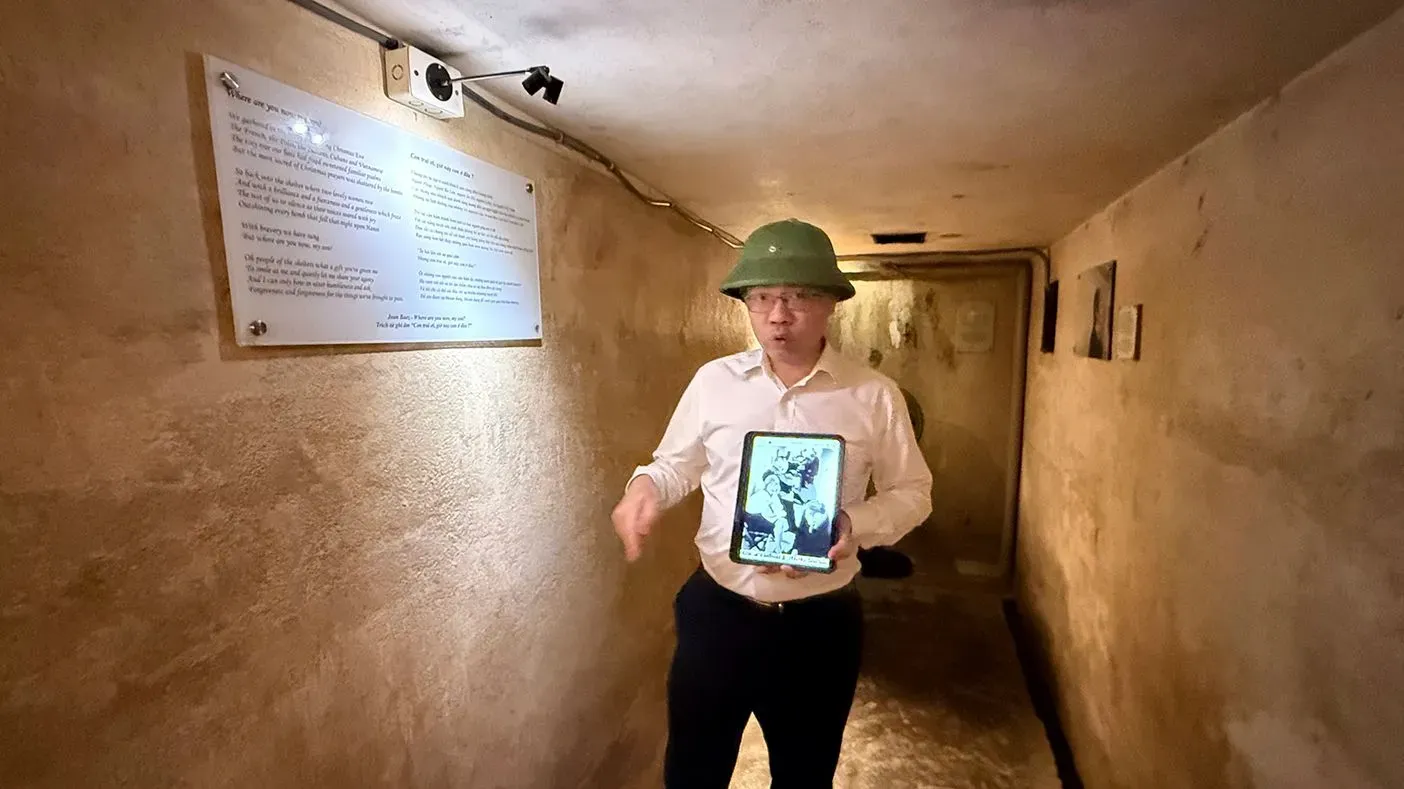 مرشد سياحي يرتدي خوذة في مخبأ تحت فندق متروبول هانوي، يعرض صورة تاريخية للزوار بينما يشرح تاريخ الفندق خلال حرب فيتنام.