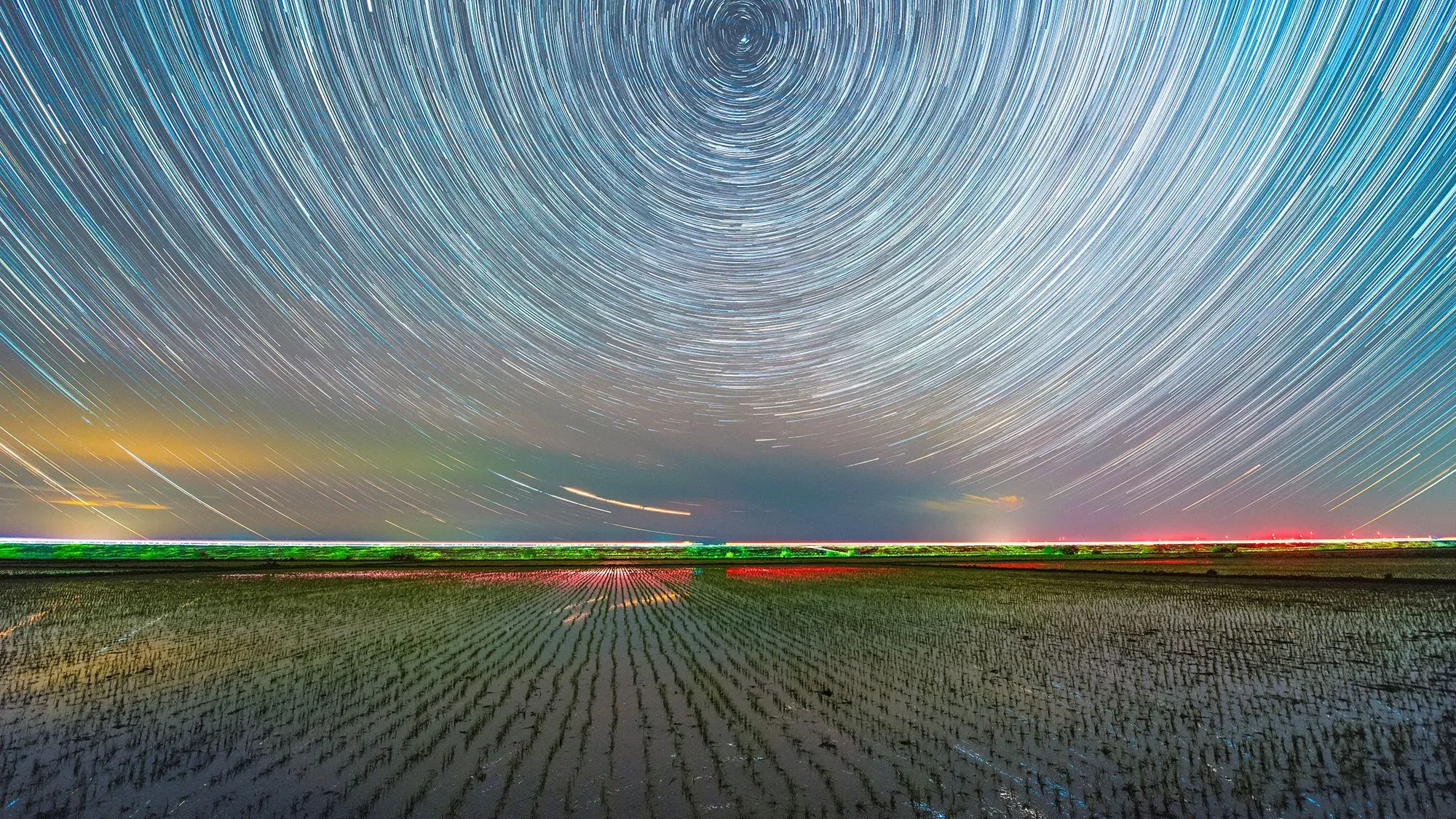 تظهر الصورة مسارات النجوم في السماء ليلاً فوق حقل زراعي، مما يبرز دوران الأرض السريع وتأثيره على طول الأيام.