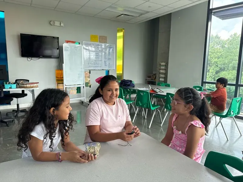 ثلاث فتيات يجلسن حول طاولة في مركز تعليمي، يتحدثن معًا بينما يظهر طفل آخر في الخلفية. يبرز أهمية البرامج التعليمية الصيفية.