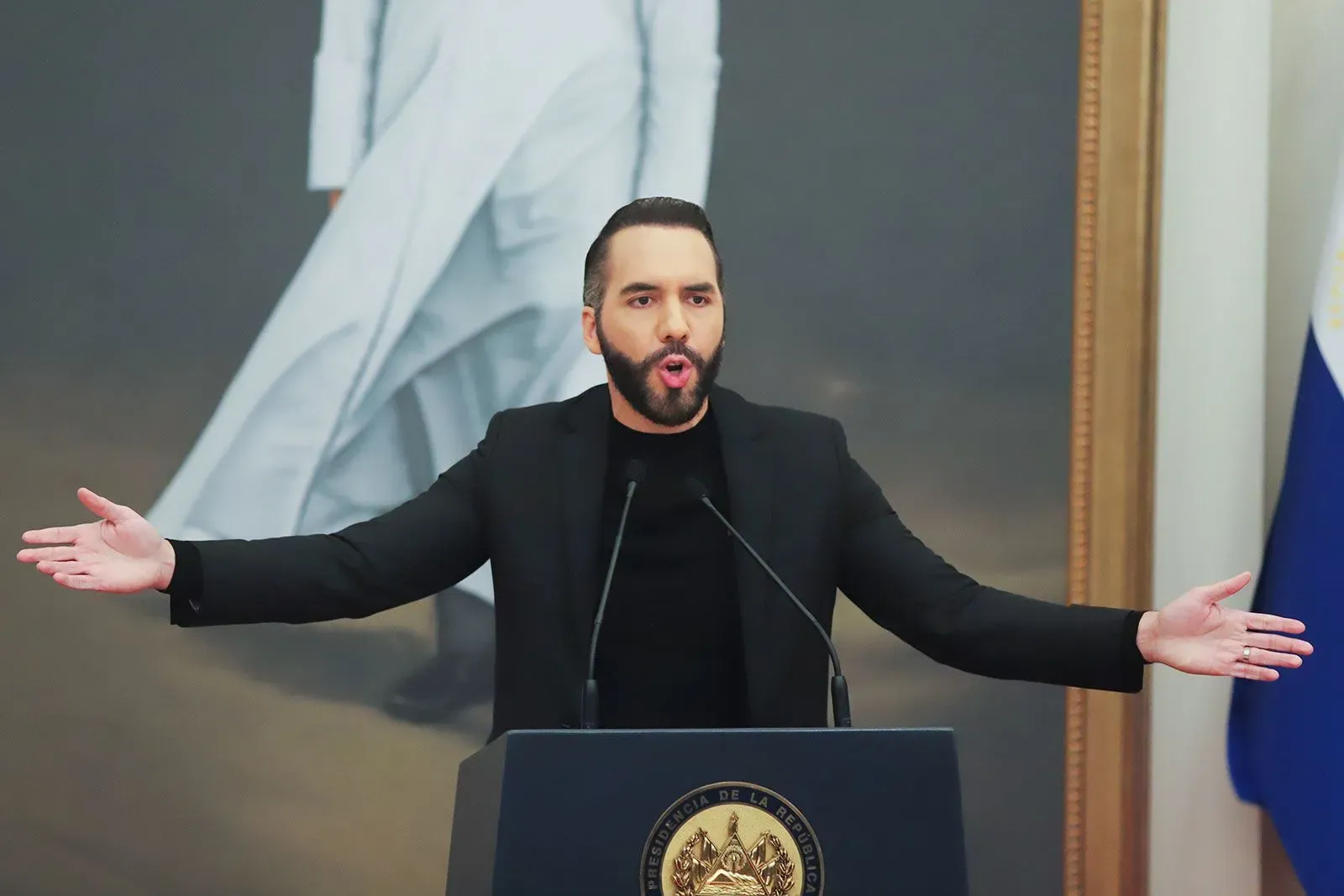 نايب بوكيلي، رئيس السلفادور، يتحدث بحماس خلال مؤتمر صحفي، مع علم السلفادور في الخلفية، وسط جدل حول تعديل الدستور.