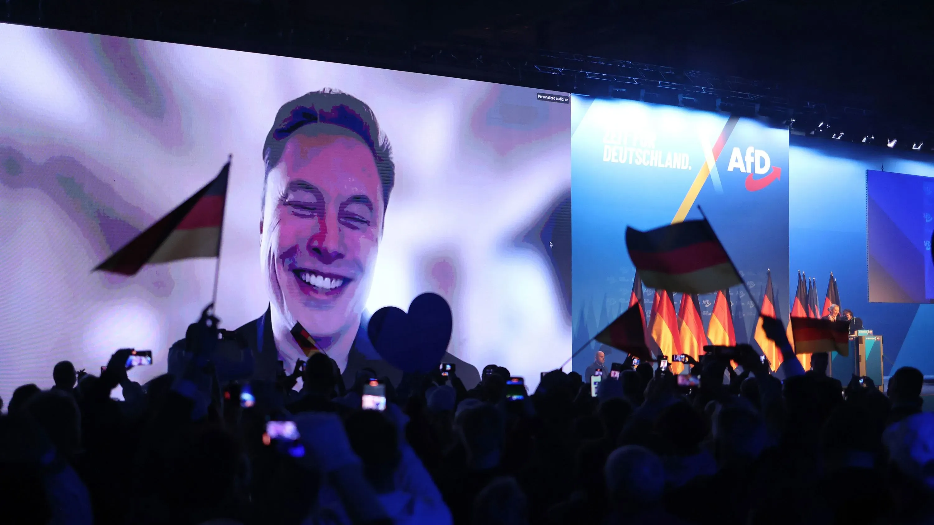 ماسك يتحدث عبر الفيديو لحشد من مؤيدي حزب البديل من أجل ألمانيا، مع رفع الأعلام الألمانية في قاعة داخلية.