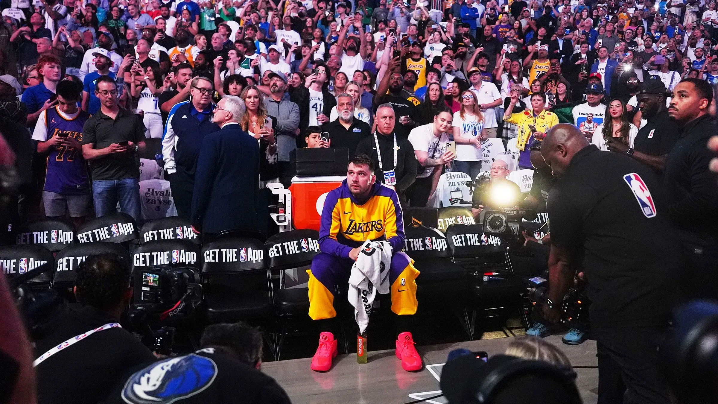 لوكا دونشيتش يجلس على مقاعد البدلاء في مباراة ليكرز ضد مافريكس، محاطًا بجمهور متحمس يهتف له بعد أدائه المذهل.