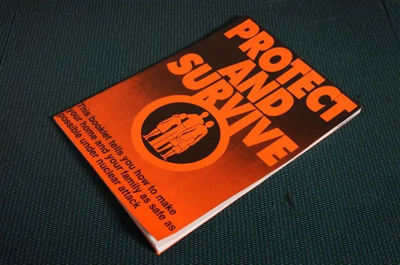 كتيب "احمِ وابقَ على قيد الحياة" يتضمن إرشادات حول كيفية تأمين المنزل والعائلة أثناء الهجمات النووية، مع تصميم بارز باللونين البرتقالي والأسود.