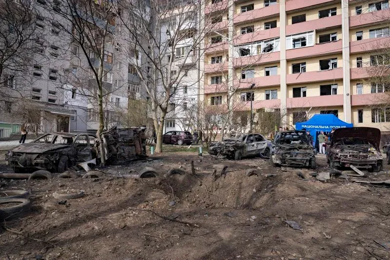 موقع حطام سيارات محترقة أمام مبنى سكني تضرر نتيجة النزاع في أوكرانيا، مع وجود أشخاص يتواجدون في المنطقة.