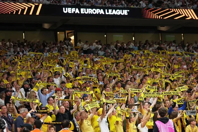 حشود جماهير نادي بودو/غليمت في ملعب توتنهام، يلوحون بأوشحة صفراء ويشجعون بفخر خلال مباراة نصف نهائي الدوري الأوروبي.